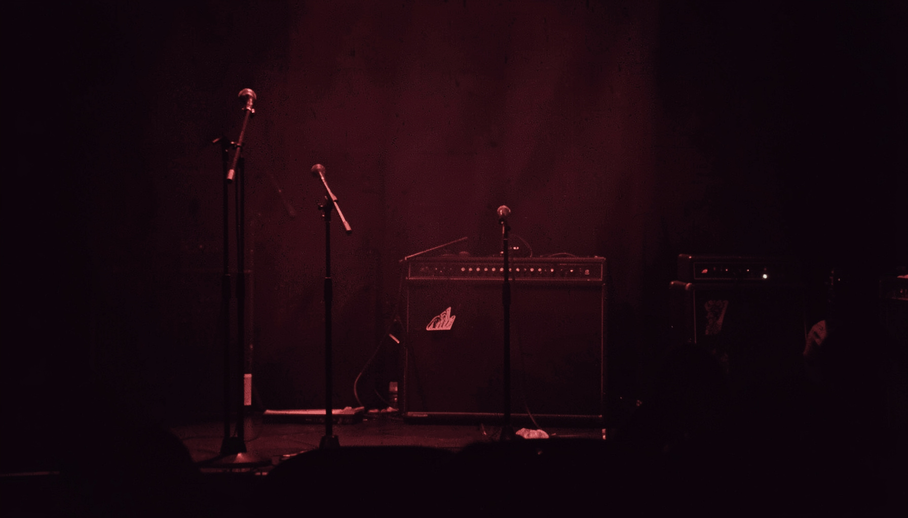 A warm duotone photograph of a dimly lit stage with microphone stands and amplifier silhouettes, deep red and charcoal tones, moody and atmospheric